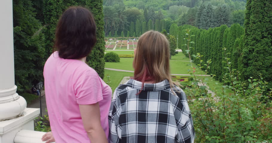 Woman in pink t-shirt and teenage girl in black white plaid shirt standing at white stone balustrade viewing manicured gardens with green lawns, hedges, flower beds, and forest backdrop.