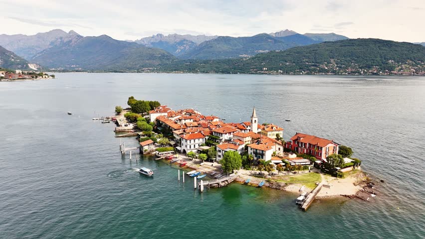 A peaceful lakeside village in Sresa on Isola Bella in Italy appears surrounded by calm water, red-roofed buildings, boats, and distant mountains creating a serene and picturesque landscape from above