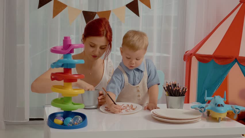 A young boy joyfully draws with his mother, using colored pencils. They are in a bright room filled with toys and vibrant decorations, enjoying quality time together.