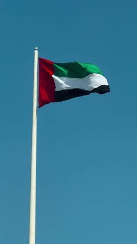 A close-up shot of the UAE national flag waving boldly atop a tall flagpole, set against a clear blue sky near Abu Dhabi, capturing national pride, color, and movement in a bright, serene atmosphere.