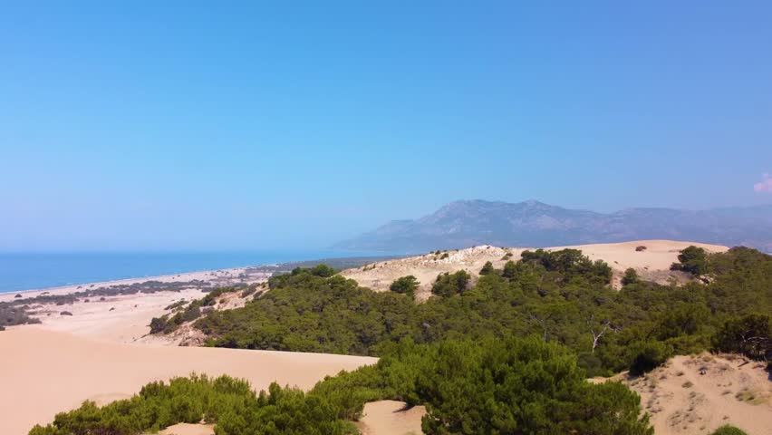 aerial drone footage capturing the vast, golden Patara Sand Dunes meeting the turquoise Mediterranean Sea in Turkey. This panoramic view showcases the unique desert-like landscape of the dunes, the ex