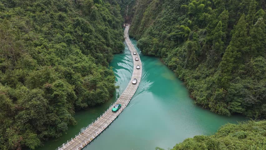Revealing aerial view of the Shiziguan floating bridge in Hubei, China, showing cars crossing over green river water surrounded by dense forest and mountains.