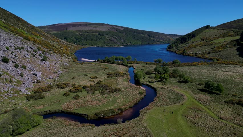 Aerial forward-moving drone view of a winding river flowing into Lough Dan, surrounded by hills and open countryside in Wicklow, Eire, capturing natural beauty in bright daylight