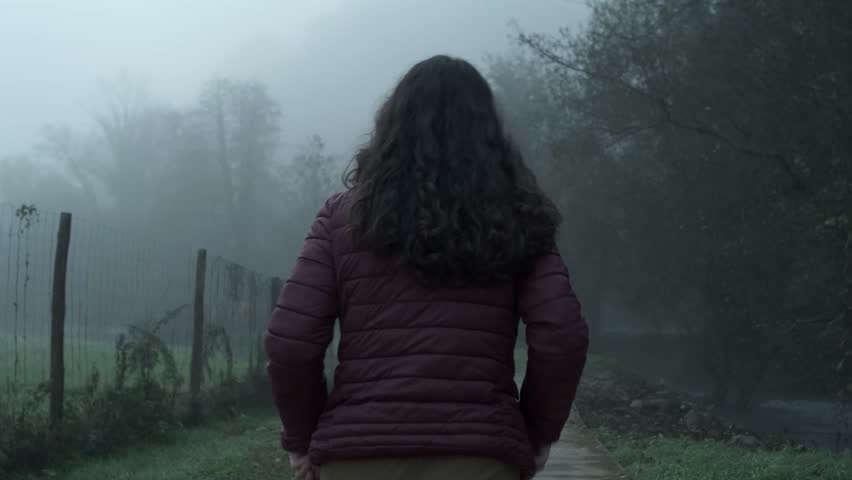 A suspenseful follow shot of a lone woman walking a dark, misty path. This atmospheric clip is perfect for creating a sense of mystery, fear, or a journey into the unknown