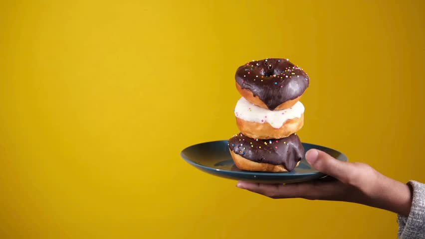 Hand holds a stack of three glazed donuts on a plate. The donuts are chocolate frosted with sprinkles and vanilla frosted. Bright Yellow background makes the donuts stand out.