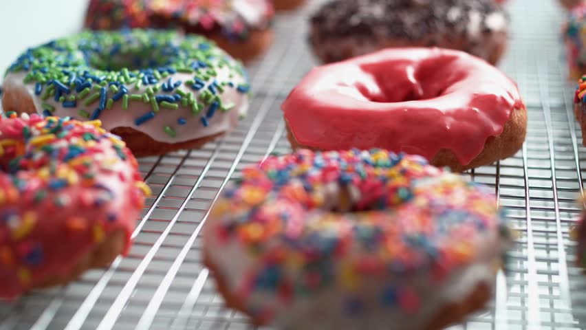 Close up of Glazed donuts are arranged on a wire rack. Pink, chocolate, and white frosted donuts are topped with rainbow and solid color sprinkles. Close up shot of sweet food.