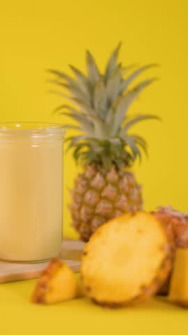 Hand places pineapple slice on creamy smoothie glass, surrounded by fresh fruit, bright yellow backdrop