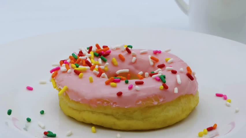 Close-up of a donut with pink frosting on a white plate. Colorful sprinkles are being dropped on the donut. White mug out of focus in the background.