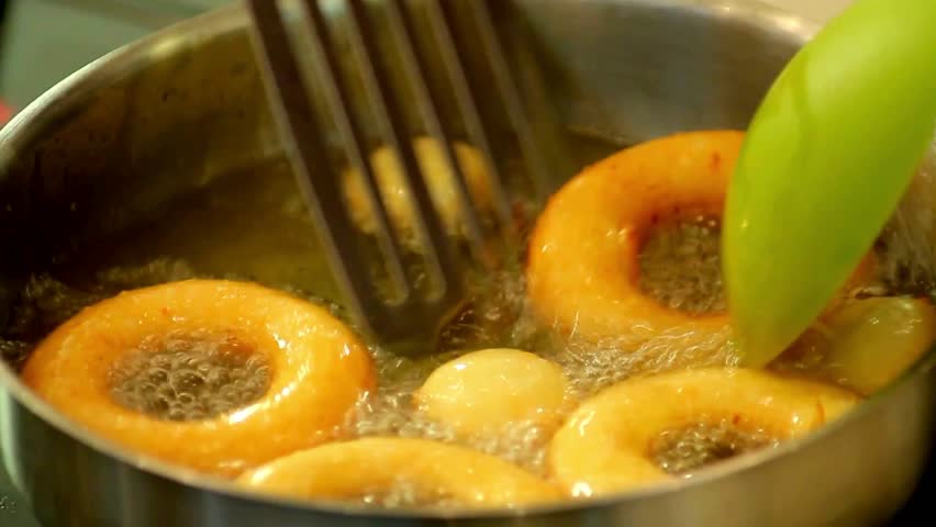 A close-up view of donuts and dough balls being fried in a hot oil in a metal cooking pot. Bubbles form around the dough as it cooks in the hot oil.