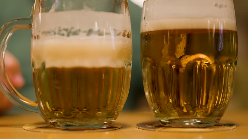 Person sets foamy lager mug on wooden table, warm indoor lighting, shallow depth of field