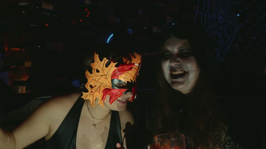 Two friends immersing themselves in the halloween spirit, one wearing a leaf mask and the other witch like makeup, enjoying a spooky night party with drinks