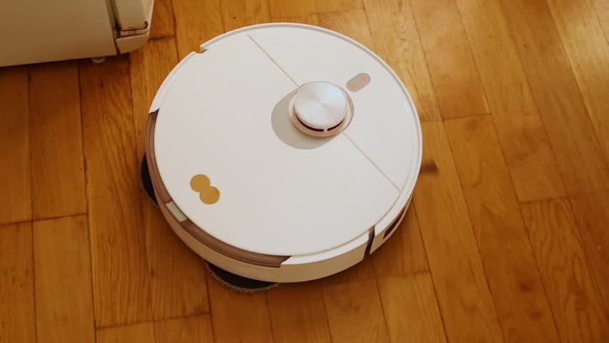 Top-down view of a white robotic vacuum cleaner moving automatically along a wall on a polished wooden floor. Highlights the lidar sensor and smart home technology for efficient, automated cleaning.
