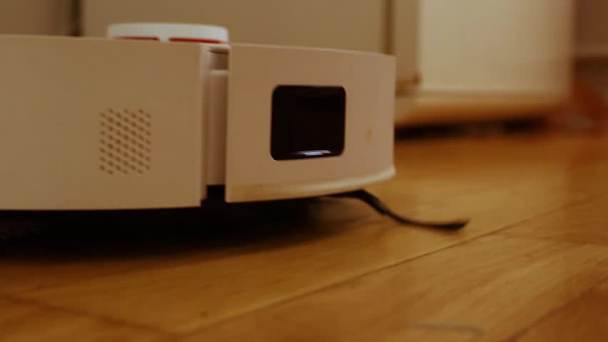Close-up view of a white robotic vacuum cleaner navigating a polished wooden floor. Focus on the lidar sensor window, side vent, and brush, highlighting smart home automation and cleaning technology.