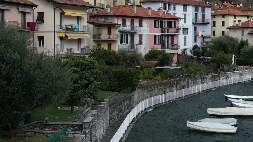 Drones capture this stunning scene of vibrant buildings alongside a tranquil lake in the Italian Alps. Boats rest gently on the water under a serene sky.