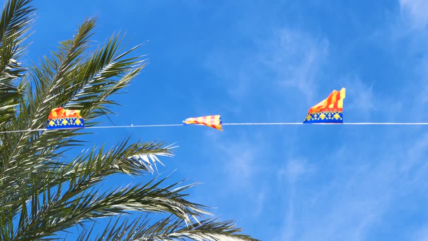 Slow motion video of Valencian flags garland fluttering among palm branches against clear blue sky, perfect for festive, travel content, summer, celebration and cultural projects