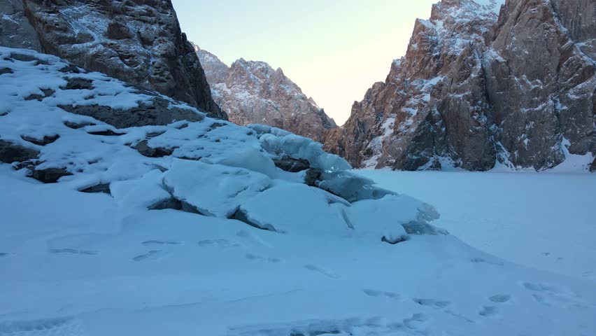 A dramatic view of the frozen Kel Suu lake, showing massive blue-tinged ice formations in the foreground and towering, sheer canyon rock walls in the background