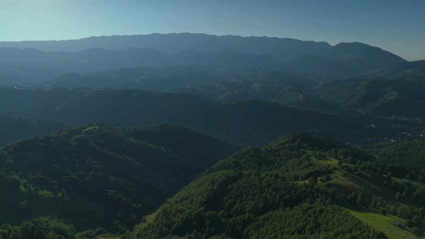 Atmospheric aerial descent over Bran Rucar corridor with Piatra Craiului Mountains fading in haze above birch-covered hills in late afternoon light