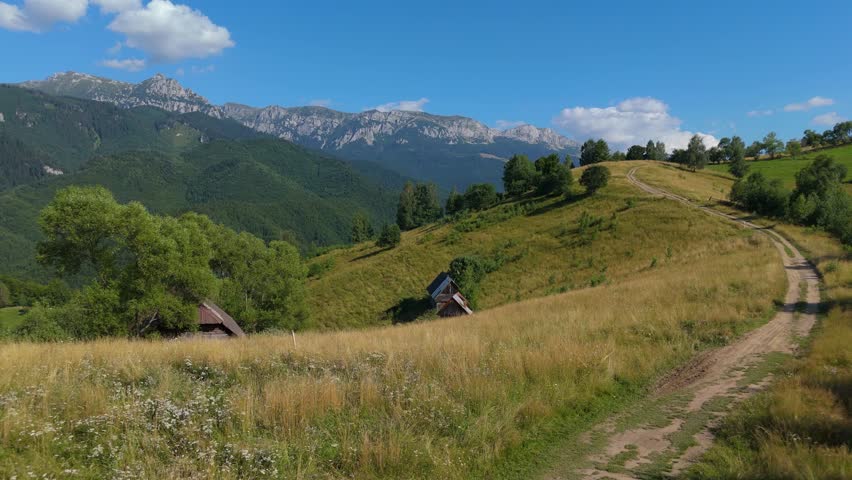 Static aerial shot of lone hiker walking away along a dirt cart road on Carpathian hills near Simon, with Bucegi Mountains ridge in the summer background