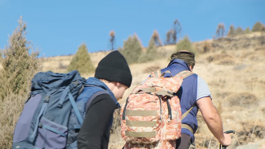 Close rear view of two hikers walking uphill one after another. An older and a younger tourist climb the slope tired and struggling. Exhausted father and son in hiking. endurance training.