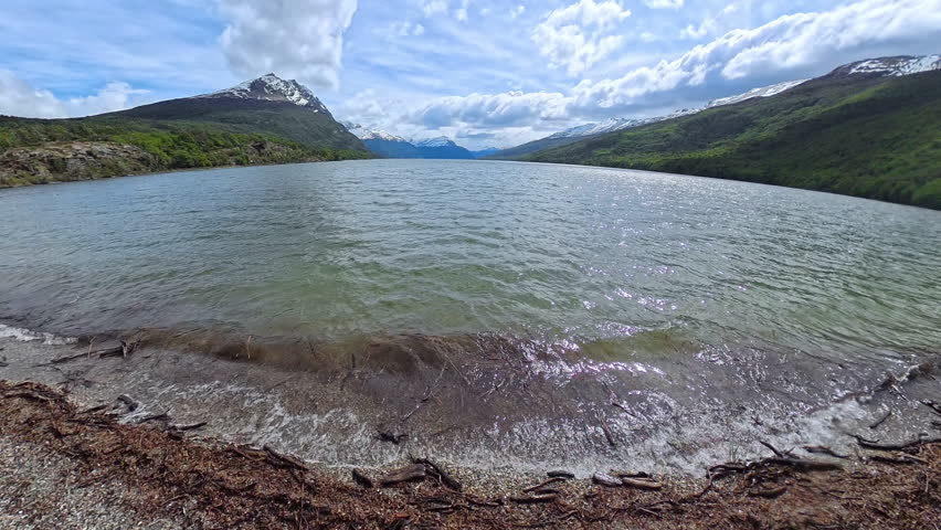 Peaceful lake in Parque Nacional Tierra del Fuego, Argentina, with small waves lapping the rocky shore. In the distance, majestic mountains rise under a calm sky, creating a tranquil landscape.