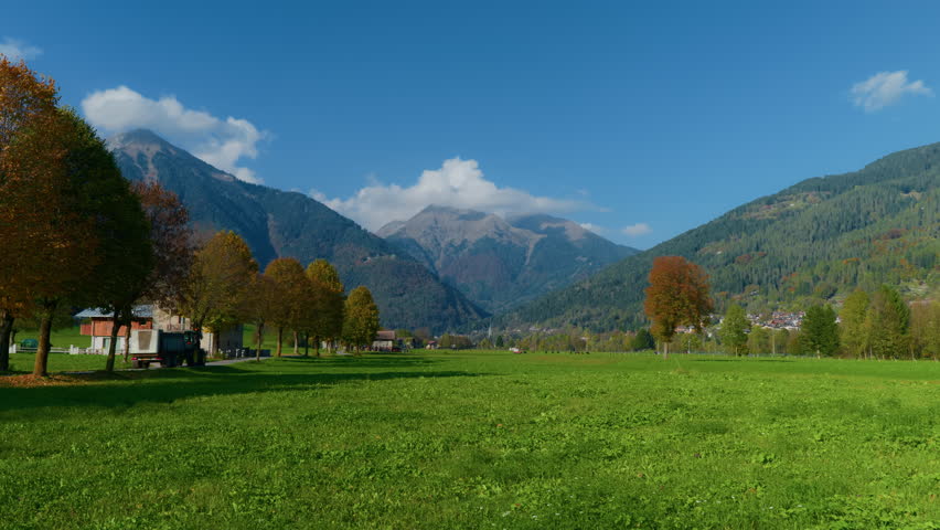Left-to-right wide shot of a mountain valley featuring barns, farmhouses, and livestock, with a tree-lined road and a tractor passing, set against distant mountain peaks.