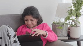 Medium shot of a preteen girl in pajamas laughing while using a tablet on a sofa. Bright window daylight with indoor plants and a lamp in a cozy home interior. - Powered by Shutterstock - Get 15% off with code: PIKWIZARD15