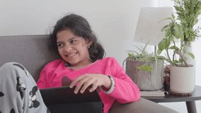 Medium shot of a preteen girl in pajamas laughing while using a tablet on a sofa. Bright window daylight with indoor plants and a lamp in a cozy home interior. - Powered by Shutterstock - Get 15% off with code: PIKWIZARD15