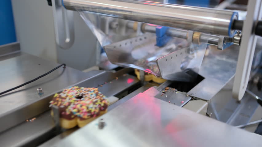 Industrial food production line wrapping fresh donuts with chocolate frosting in plastic packaging