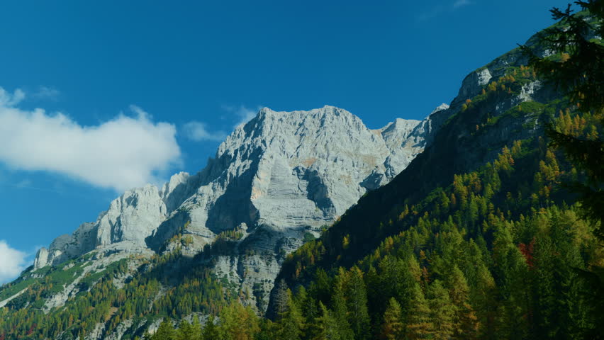 Pan shot from left to right showing a rocky mountain peak with a lush forested valley below, highlighting the natural beauty and rugged alpine landscape.