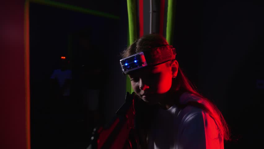 Portrait of an active girl enthusiastically playing laser tag in a dark room with neon stripes on the wall. A girl with a laser gun and a marker on her head is playing laser tag.