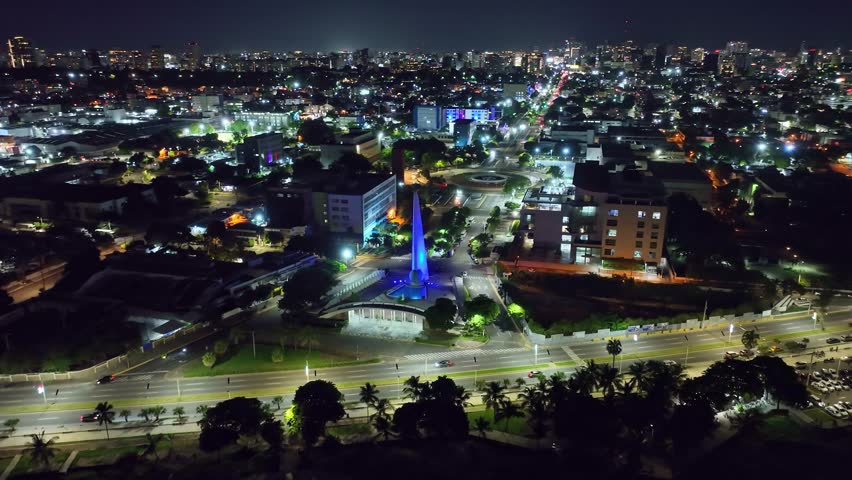 Aerial drone view of the Centro de los Héroes district in Santo Domingo at night, illuminated monument in a roundabout, city lights, traffic, and the urban skyline of the Dominican Republic