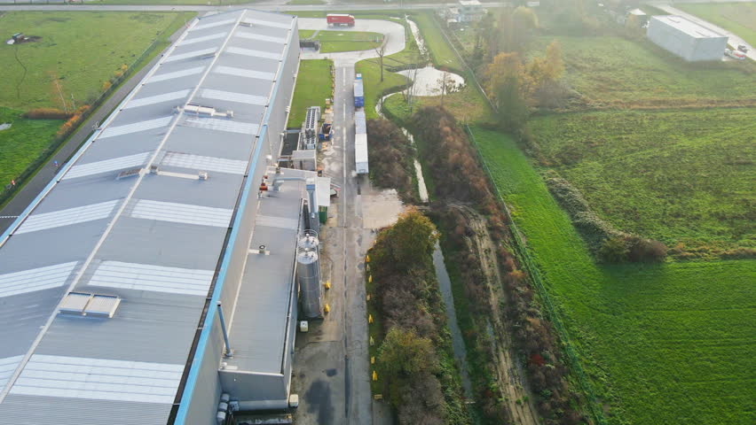Aerial of a large factory with a red truck parked in front of the warehouse. The industrial site is surrounded with green meadows while the sun shines brightly.