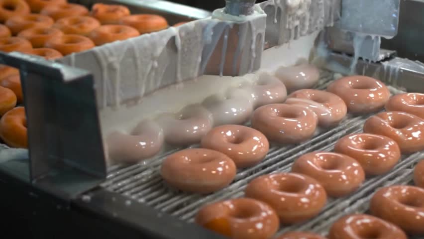Donut being glazed on conveyor belt in factory Fresh, donuts travel on conveyor belt getting glazed with white icing in a donut factory. Bakery products in process. Food industry. close up, 4k uhd