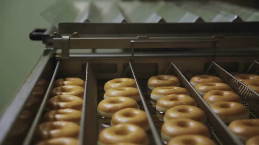 Eye-level view of several donuts being produced on a conveyor belt in a food factory process background. Donuts slide along a production line.