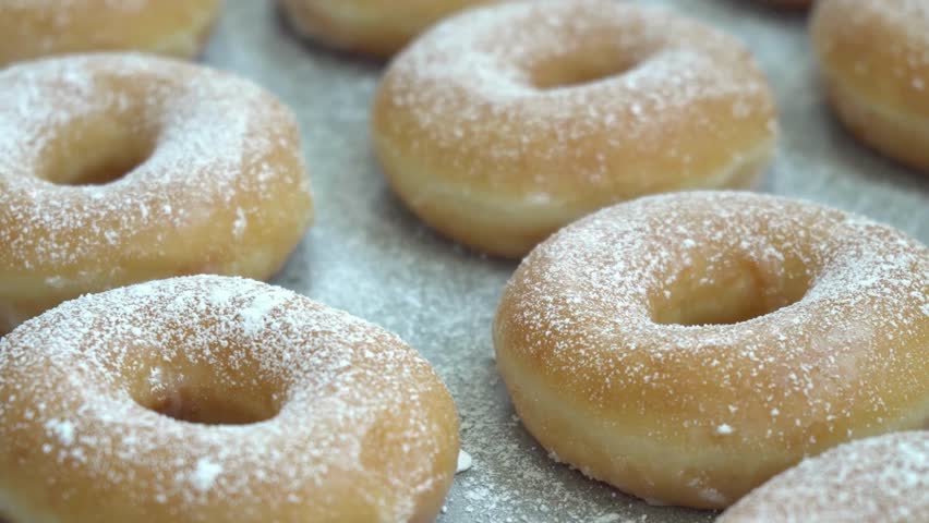 Donuts covered with powdered sugar sit on a tray, freshly prepared. These glazed donuts are ready to be eaten. Close up perspective.