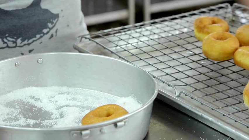 Freshly made donuts are being coated with granulated sugar in a stainless steel pan. Several glazed donuts sit cooling on a wire rack nearby.
