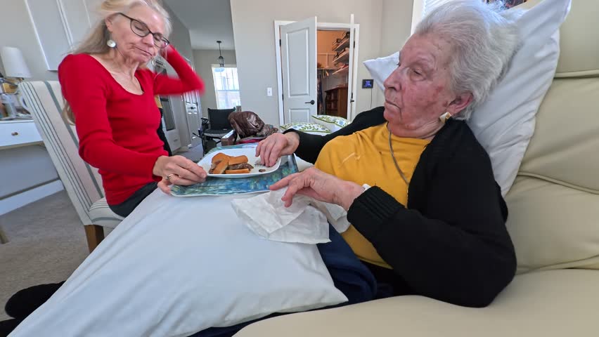 An elderly woman receives loving attention from her daughter during a nursing home visit as they share a snack and conversation.