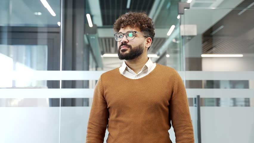 Portrait of smiling businessman standing in modern glass business office. Happy successful male professional in casual sweater and glasses looking at camera. Handsome manager posing with crossed arms