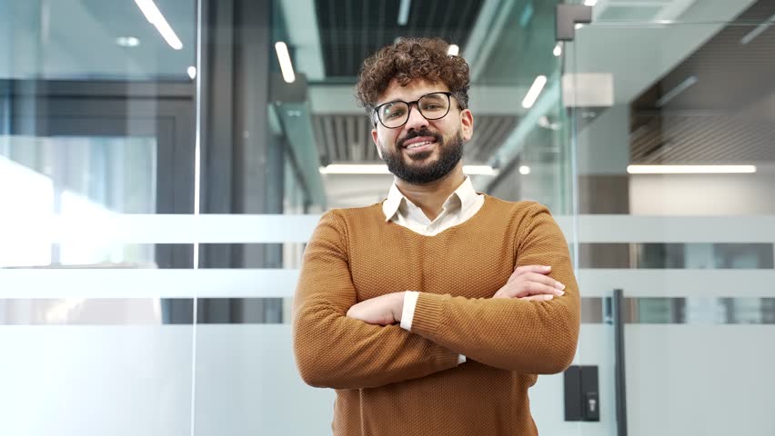 Portrait of smiling businessman standing in modern glass business office. Happy successful male professional in casual sweater and glasses looking at camera. Handsome manager posing with crossed arms