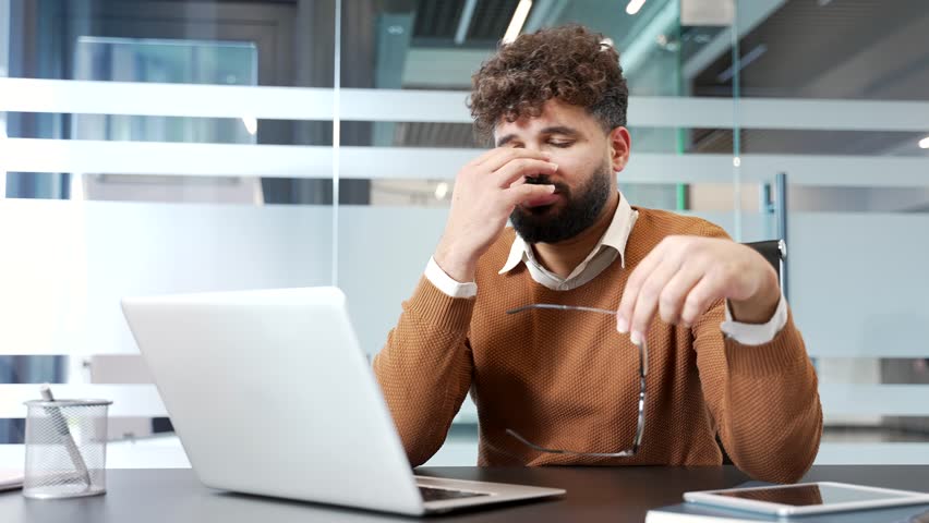 Businessman feels eye fatigue while working on a computer sitting at workplace in business office. Overworked man suffering from eye strain. Worker takes off glasses and massages, rubs bridge of nose