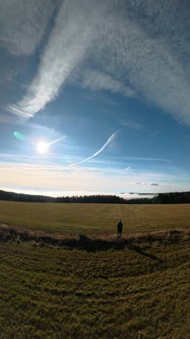 A lone person stands in a wide sunlit field beneath dramatic cloud streaks in a blue sky