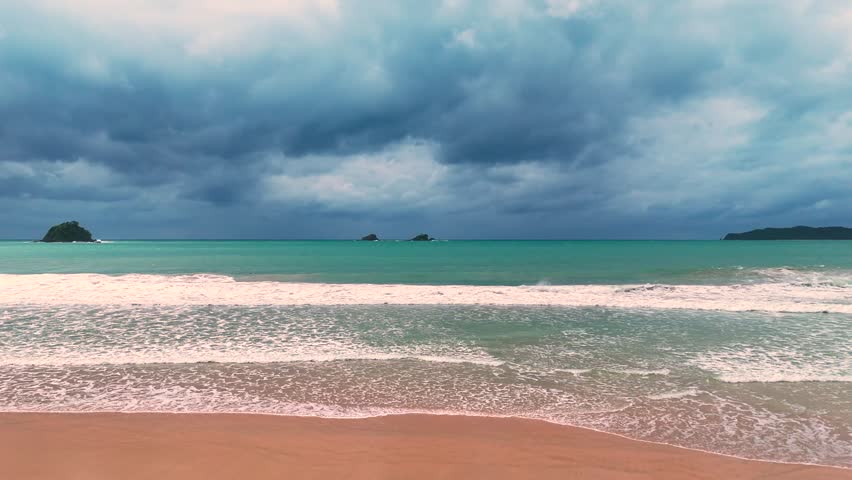 Tranquil view of a tropical beach with turquoise waters gently lapping against the shore, framed by dramatic, cloudy skies in the background.