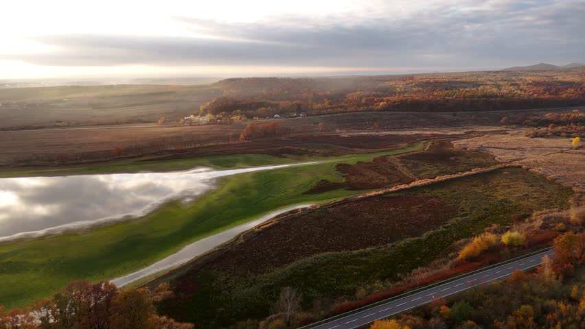 A calm autumn landscape with an old lake reflecting the sky, surrounded by fields and colorful trees. A quiet road runs along the edge, adding a peaceful rural atmosphere.