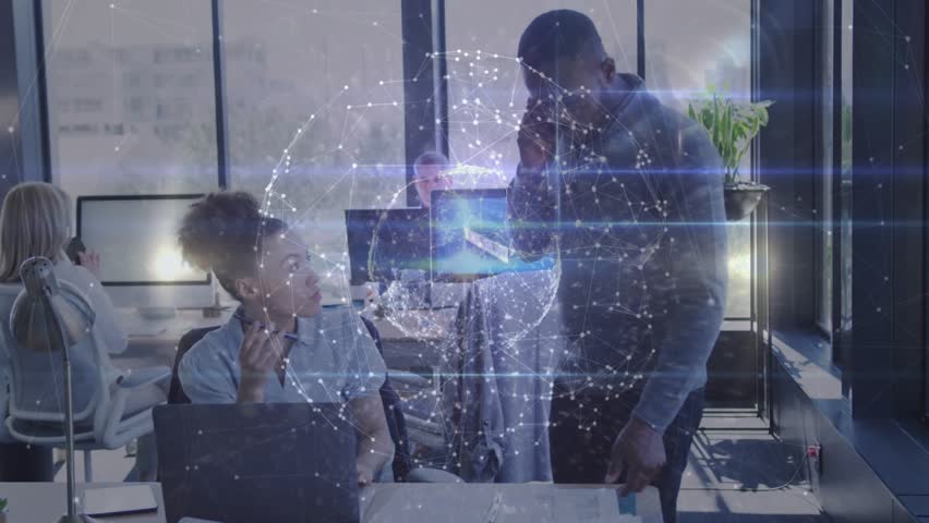 Seated woman, standing man ending call, arranging papers on laptop while globe pulsing in business. Collaboration, desk, office, openplan, daylight, monitors, documents