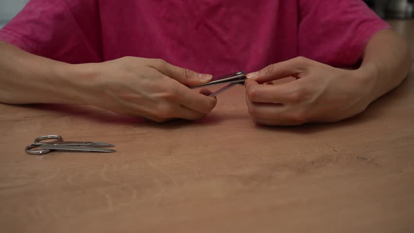 Woman in a pink shirt sitting at a wooden table, carefully trimming her fingernails with metal nail clippers, focusing on personal hand care and hygiene with a pair of scissors nearby