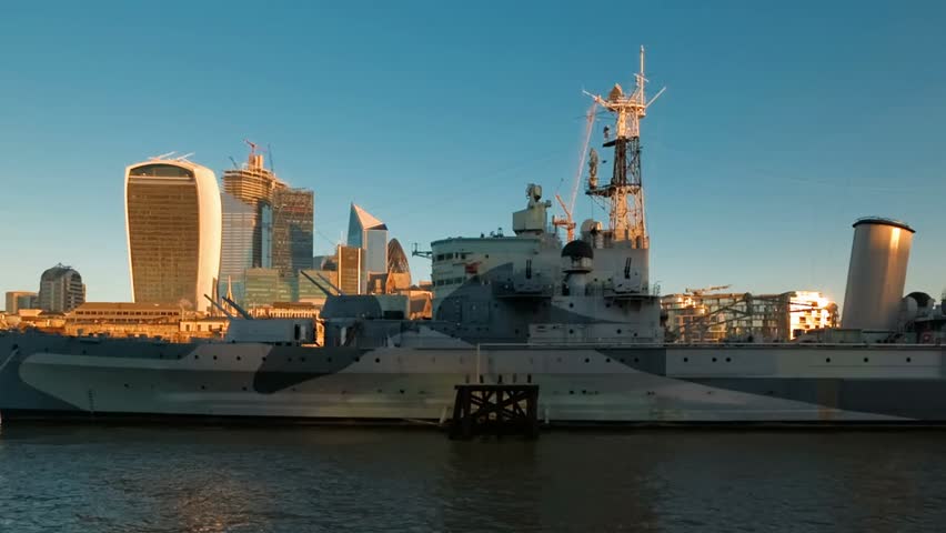 Wide cityscape view featuring a historic naval cruiser on a calm river with modern skyscrapers rising behind it, blending maritime heritage with urban architecture.