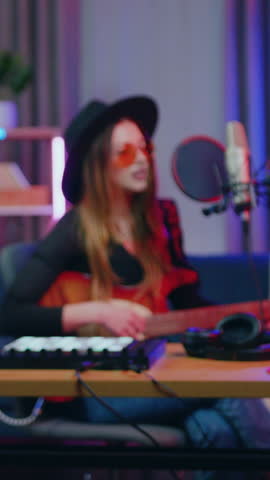 Woman singer in hat and glasses pushing buttons on drum midi player starting to sing song into recording equipment to the sounds of her guitar in home studio