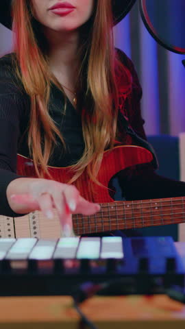 Lovely positive successful 25-aged long -haired girl in black hat and sunglasses recording new song with special drum machine and guitar in specially equipped place at home with night lightings