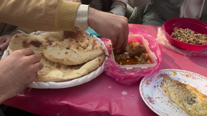 Close-up 4K slow-motion of people sharing a traditional Pakistani breakfast outdoors, dipping naan into curry with a fried egg on the table. A warm, authentic street-food and cultural moment