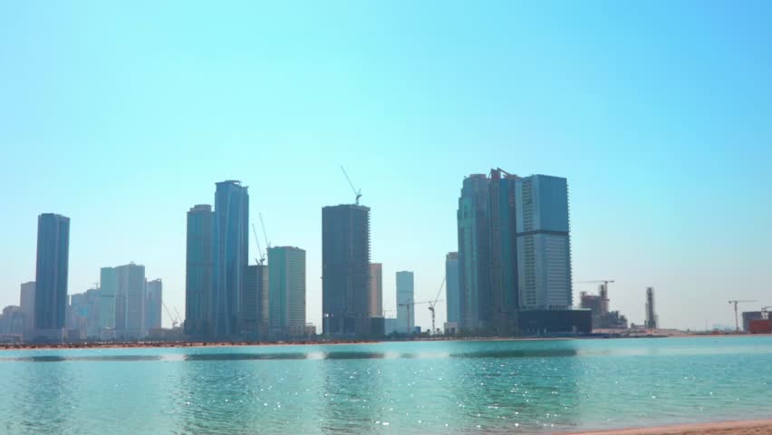 Panoramic city view of Sharjah, UAE, showcasing modern skyscrapers, coastal waterfront, and contemporary urban architecture. Clear daylight enhances the skyline, reflecting buildings in the calm water